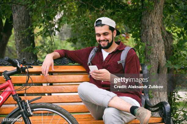 a smiling hindu young man sits on a wooden bench in a public park. a dark-skinned indian man in a baseball cap, texting and communicating on the internet social networks on a mobile phone. there is a bicycle nearby. the concept of a healthy lifestyle. - indian male model stockfoto's en -beelden