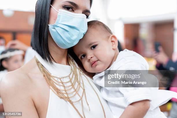 latina mom wearing mask carries her 1 year old baby both in the church where the baby's christening is celebrated - priest baptism stock pictures, royalty-free photos & images