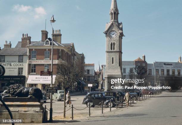 Pedestrians walk past cars parked by the Clock Tower at Market Square in the town of Aylesbury in Buckinghamshire, England circa 1960.