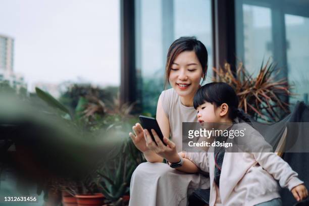 young asian mother using smartphone with lovely little daughter while relaxing on deck chair in the backyard, surrounded by beautiful houseplants. family lifestyle and technology - family holidays hotel stockfoto's en -beelden