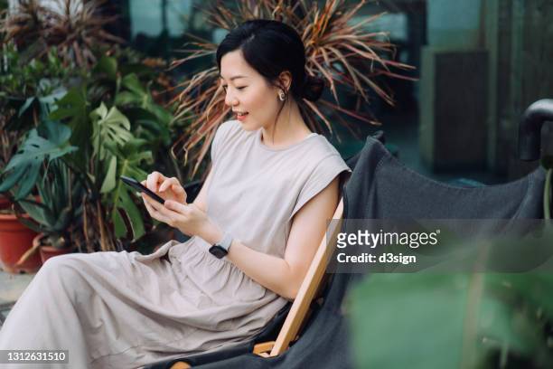 beautiful young asian woman using smartphone while relaxing on deck chair in the backyard, surrounded by beautiful houseplants. lifestyle and technology - asian using phone stock pictures, royalty-free photos & images