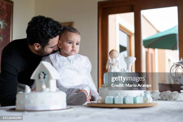 de vader let op zijn baby aangezien hij bij de lijst naast de doopkoeken zit terwijl de baby zijn doopjurk draagt - dopen stockfoto's en -beelden