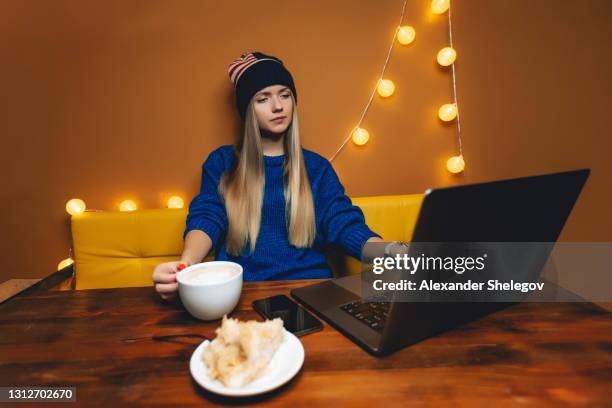 young woman is drinking coffee indoors. female portrait with cup of hot drink. girl is eating sweet cake and enjoying by work online on laptop computer. freelance concept - orange hat stock pictures, royalty-free photos & images