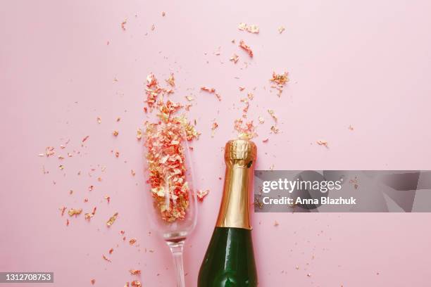 green and golden glass bottle of champagne, glass and golden confetti over pink background. festive new year greeting card. - exploding glass stockfoto's en -beelden