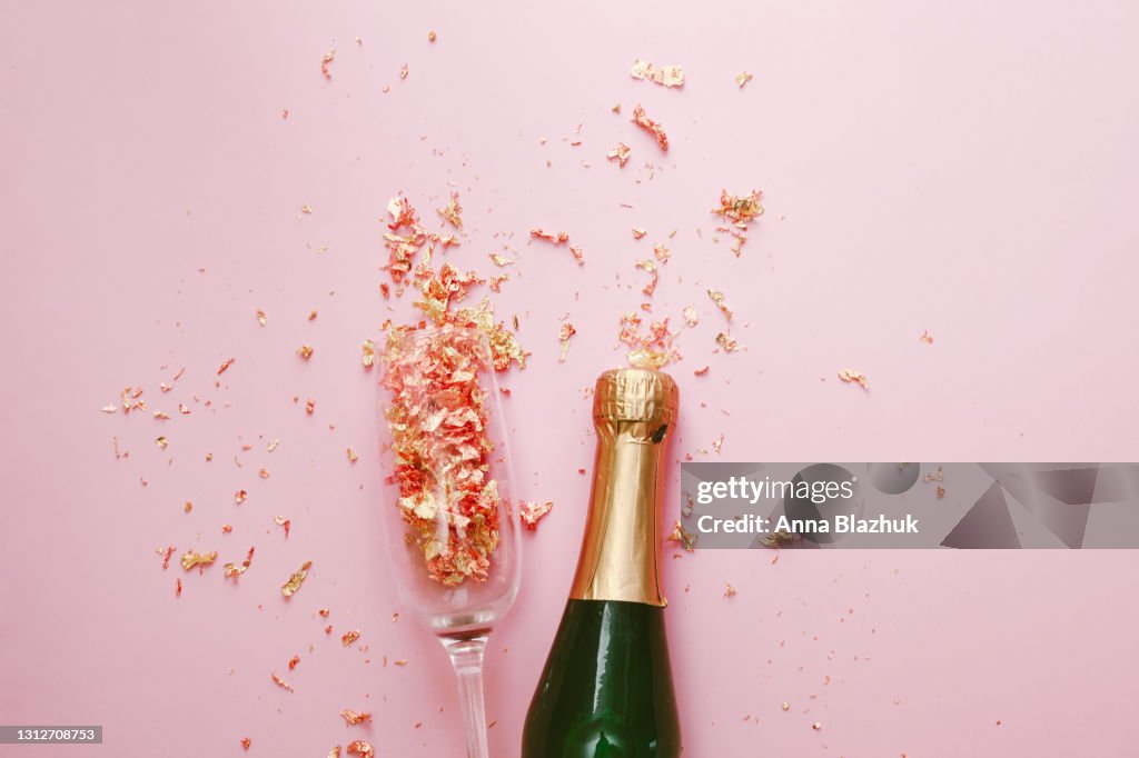 Green and golden glass bottle of champagne, glass and golden confetti over pink background. Festive New Year Greeting Card.