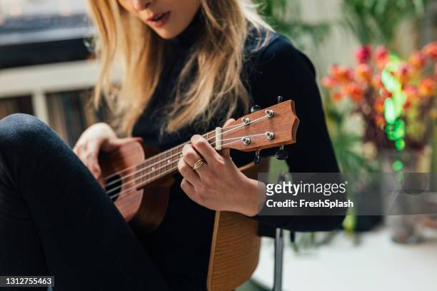 young beautiful caucasian female artist sitting in her studio apartment and playing ukulele - songwriter stock pictures, royalty-free photos & images
