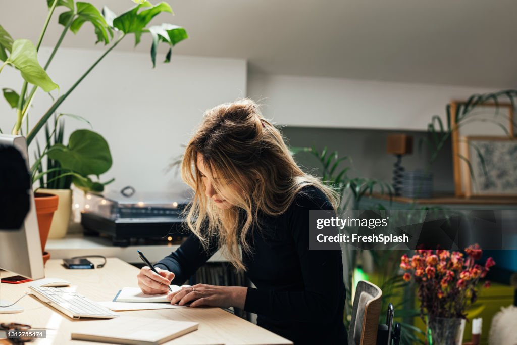 Junge Kaukasierin, die von zu Hause aus arbeitet- in ihrem Bürobereich sitzt und Notizen schreibt (Horizontal)