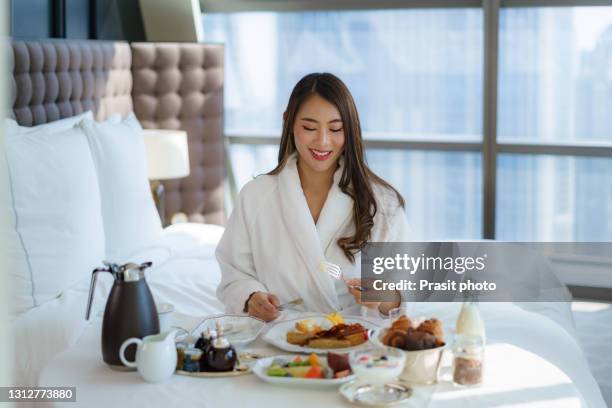 asian woman in pajamas eats breakfast in bed in a bedroom in a luxury downtown hotel during a vacation trip. - hotel de lujo fotografías e imágenes de stock