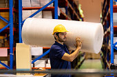 Male employee warehouse worker wearing safety helmet and blue uniform carrying roll shockproof plastic or air bubble wrap in the industry factory warehouse. Inspection quality control