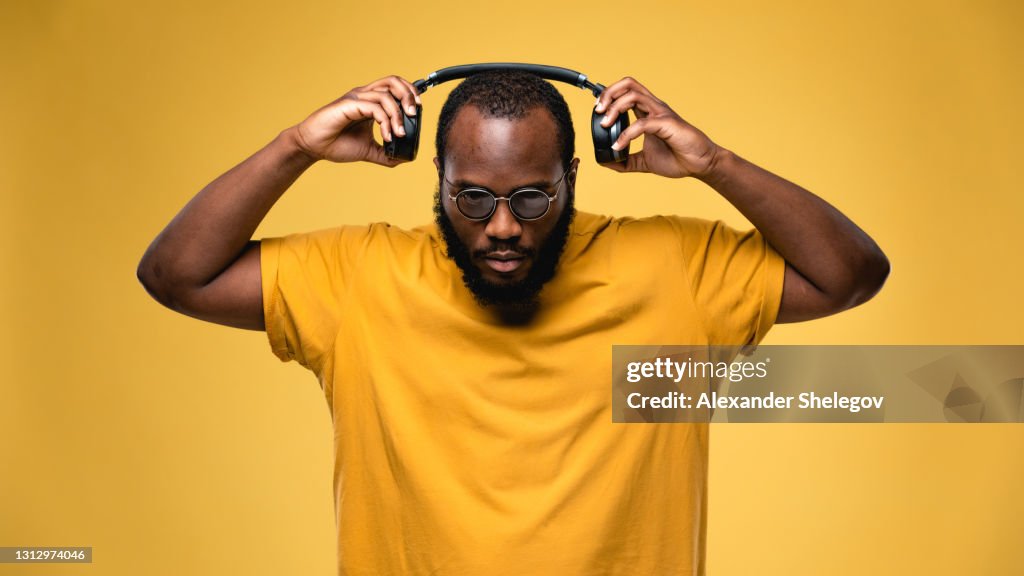African-american ethnicity person with eyeglasses and wireless black headphones is listening music. Concept for dj, techno, pop, rap, r&b music photography. Studio portrait on yellow background