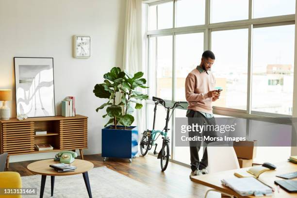 man using smart phone while leaning on window - man living room fotografías e imágenes de stock