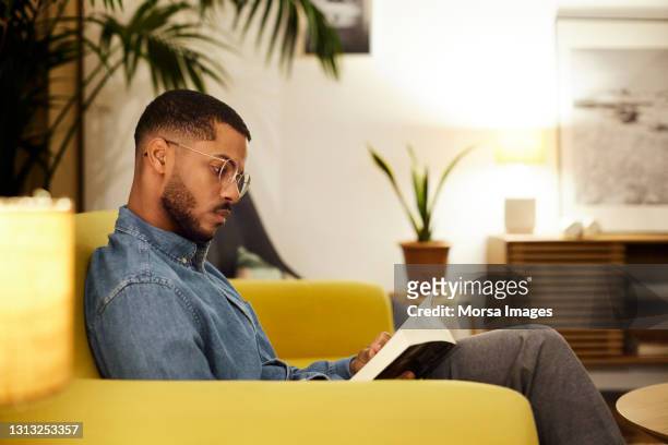 relaxed young man reading book in living room - handsome stock pictures, royalty-free photos & images