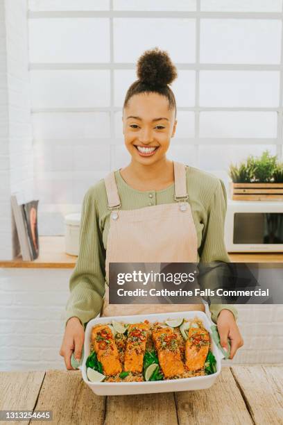 confident happy young woman holds a freshly prepared salmon meal in an oven dish - dining presentation food stock-fotos und bilder