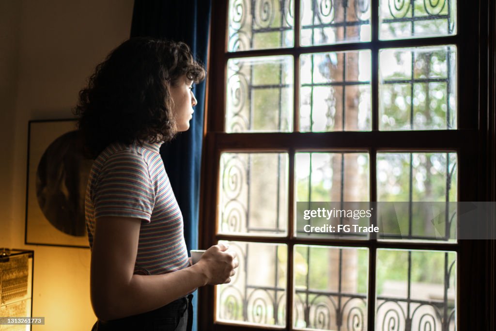 Transgender woman drinking tea looking through the window at home