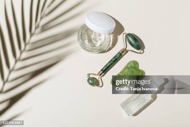 cosmetic glass bottle with pipette, moisturizing gel and jade face roller, guasha on beige background with shadow of tropical leaf. skincare beauty product. flat lay, top view, copy space. - spooning stock pictures, royalty-free photos & images