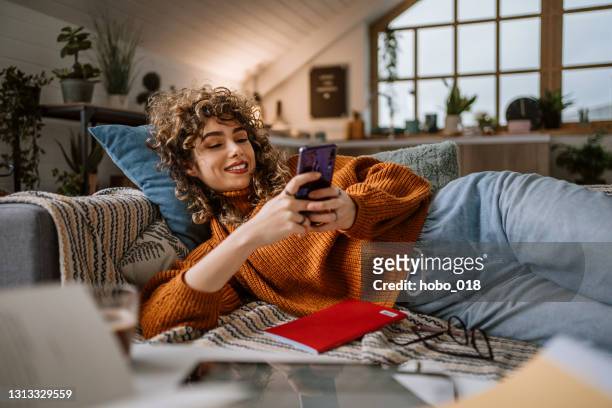 femme utilisant le téléphone intelligent pour des médias sociaux s’étendant dans son divan - femme salon photos et images de collection