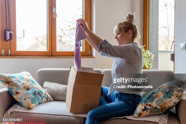 la jeune boîte en carton ouverte attirante de jeune femme heureuse satisfaite de la commande en ligne d’achat de magasin s’asseyent sur le sofa à la maison. - article de presse photos et images de collection