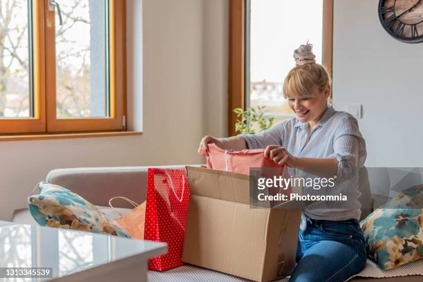 la jeune boîte en carton ouverte attirante de jeune femme heureuse satisfaite de la commande en ligne d’achat de magasin s’asseyent sur le sofa à la maison. - box in open photos et images de collection