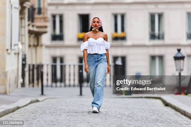 Alicia Aylies wears a red and white striped scarf over the head, a white pleated off-shoulder low-neck top with ruffle and puff sleeves, blue large...