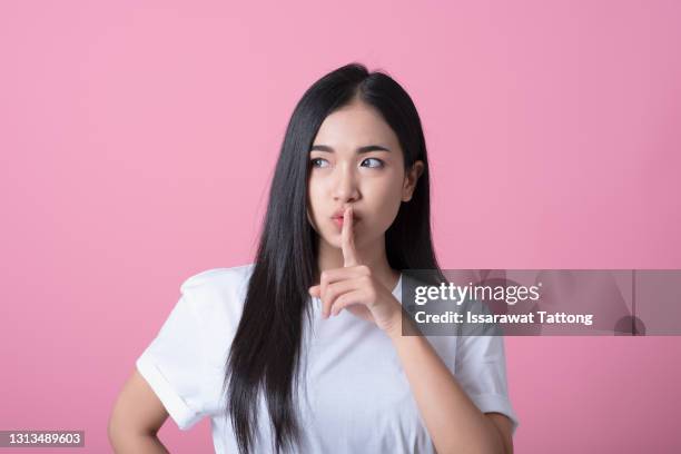 index finger on lips, silence gesture, shhh quiet, asks for voicelessness forefinger at the mouth. young attractive woman in a light t-shirt on pink background - finger on lips stock-fotos und bilder