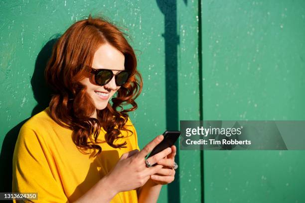 woman using smart phone, smiling, next to green wall - portraits fun mobile wall stock-fotos und bilder