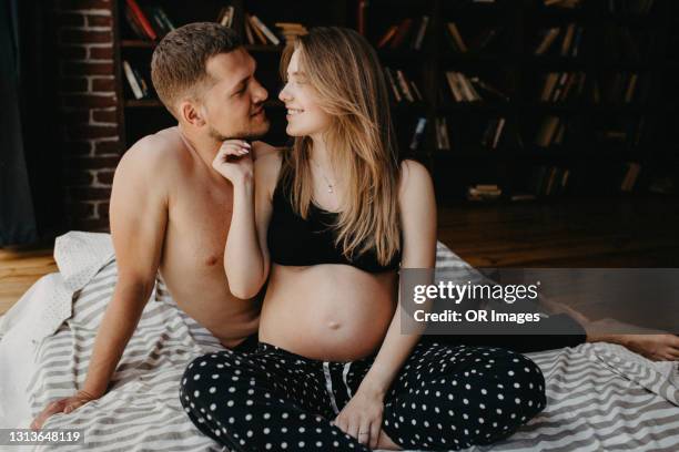 affectionate man and pregnant woman sitting on bed at home - halfgekleed stockfoto's en -beelden