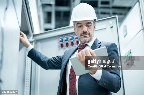 mature male electrical engineer working with digital tablet standing in front of control panel testing and record results about power distribution system in control room of factory. - energie sparen stock-fotos und bilder