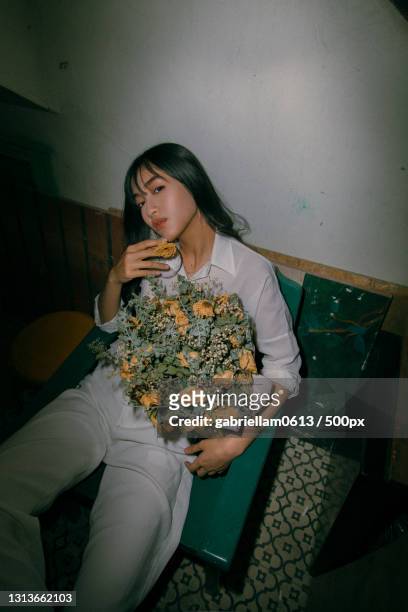 portrait of young woman holding flowers while sitting on chair at home - redactioneel stockfoto's en -beelden