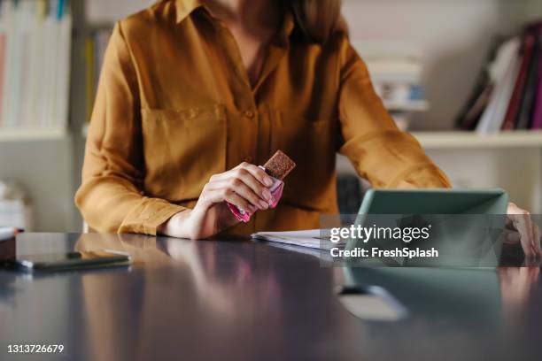 onherkenbare onderneemster die een eiwitreep op het bureau eet, terwijl het bijwonen van een videovergadering met haar collega's - staples office stockfoto's en -beelden