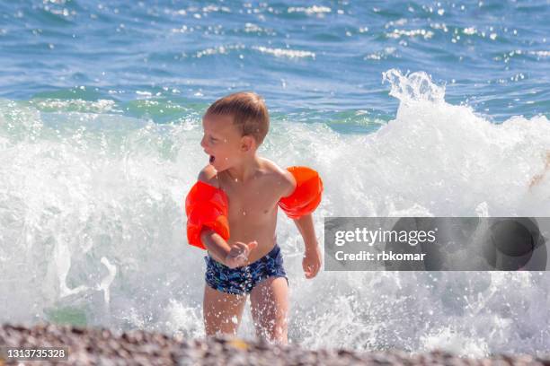 cheerful screaming toddler boy in water wings runs from a large wave on the sea beach. swimming child playing in water with splashes on summer vacation - the runaways photos et images de collection