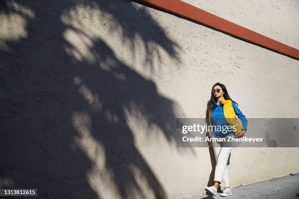 brunette woman wearing blue shirt and yellow puffer vest while walking next to the wall with palm tree shadows - city life authentic stock pictures, royalty-free photos & images