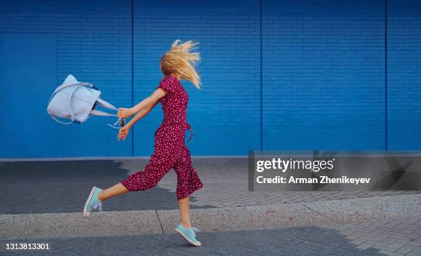 unrecognizable blond woman wearing burgundy jumpsuit and holding cerulean tote bag while jumping against the blue wall - blue jumpsuit - fotografias e filmes do acervo