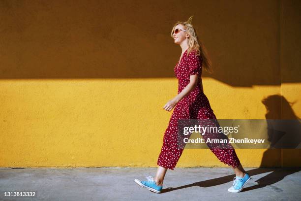 blond woman wearing burgundy jumpsuit and sunglasses while walking against the yellow wall - miami people stock pictures, royalty-free photos & images