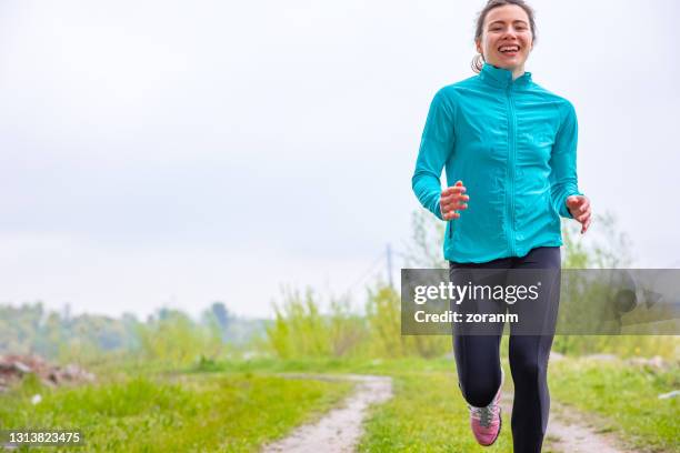 smiling teenage girl jogging on the road and looking at camera - windbreak stock pictures, royalty-free photos & images