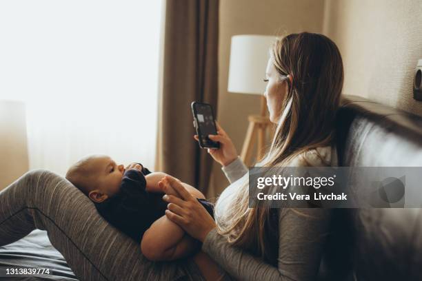 mother and baby playing with a smart phone sitting on a couch in the living room at home - eltern baby stock pictures, royalty-free photos & images