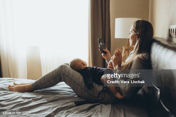 mother and baby playing with a smart phone sitting on a couch in the living room at home - eltern stock pictures, royalty-free photos & images