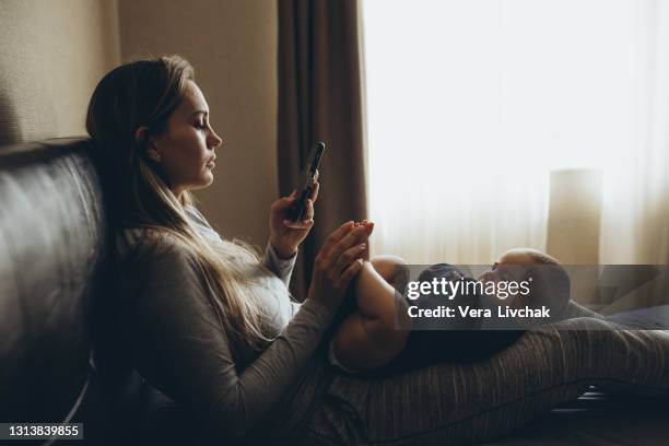 mother and baby playing with a smart phone sitting on a couch in the living room at home - eltern stock pictures, royalty-free photos & images