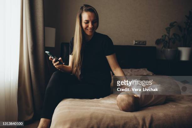mother and baby playing with a smart phone sitting on a couch in the living room at home - eltern baby stock pictures, royalty-free photos & images