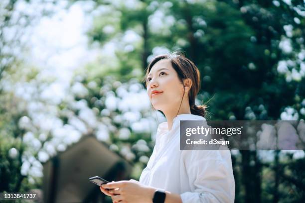 young asian woman using smartphone outdoors in the nature, against sunlight and green plants. looking up to sky with positive emotion and smile. taking a break in urban park - business park stockfoto's en -beelden