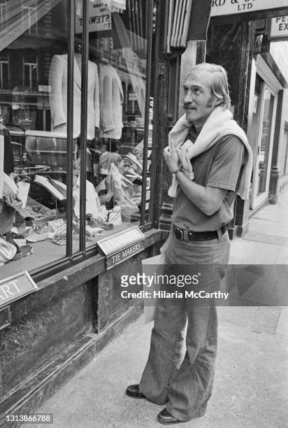 American lawn tennis player Stan Smith, with a sweater draped over his shoulders, inspects the window of an unspecified men's clothing shop in...