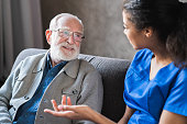 Female african professional doctor consulting senior patient grandfather during medical care visit. Young woman physician and old man talking providing medical assistance sitting on sofa. Elderly people home care concept
