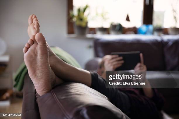 man relaxing on sofa in living room with feet up and reading on a handheld device - feet up stock pictures, royalty-free photos & images