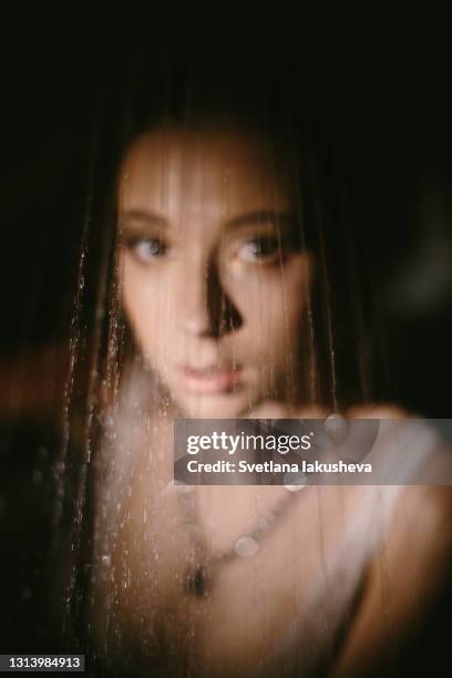 defocused portrait of young brunette woman in low key in lingerie through water stream of drops from shower - soft focus eye water stock-fotos und bilder