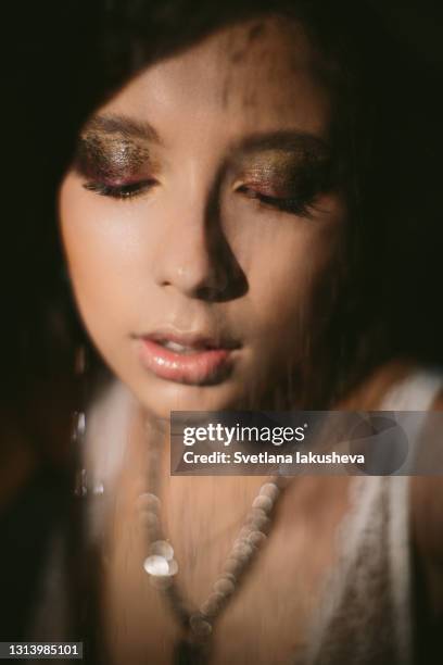 portrait of young brunette woman in low key in lingerie through water stream of drops from shower. the model looks down. - soft focus eye water stock-fotos und bilder