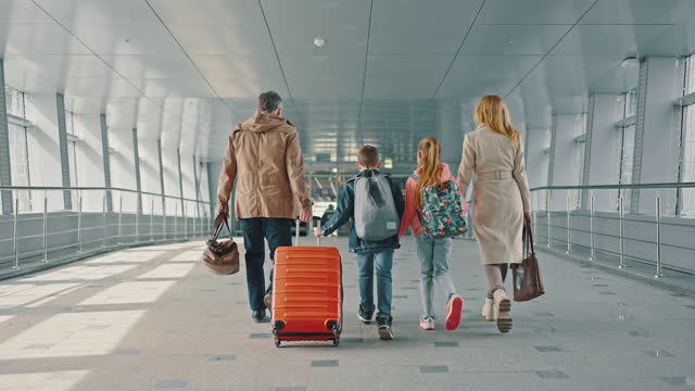 Family of two parents and two kids with luggage walking down the airport terminal, wearing protective masks, back view