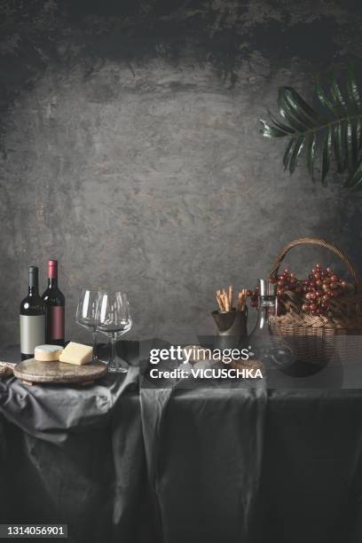 stylish still life with red wine bottles, cheese, baguette bread, snacks, grapes in wooden basket and wine glasses with carafe on table with grey tablecloth. - stillleben stock-fotos und bilder