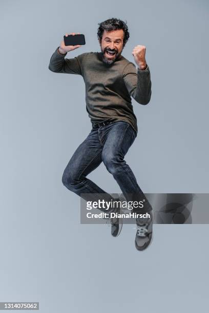 studio shot of a exited happy screaming and exited mature man with casual clothings jumping high with his mobile phone in the hand - man studio shot stock pictures, royalty-free photos & images