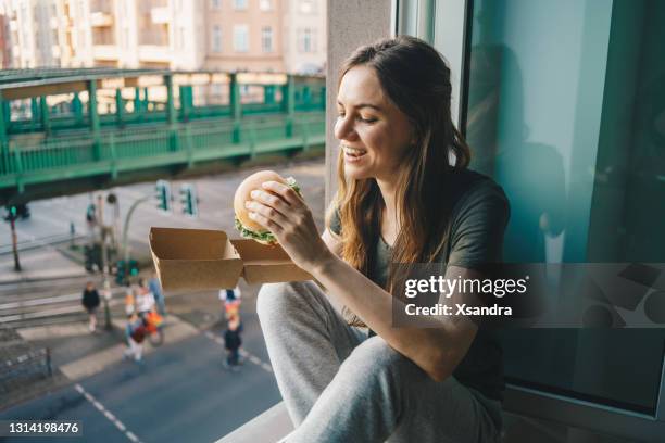 frau essen nehmen burger zu hause vor dem offenen fenster - burger stock-fotos und bilder