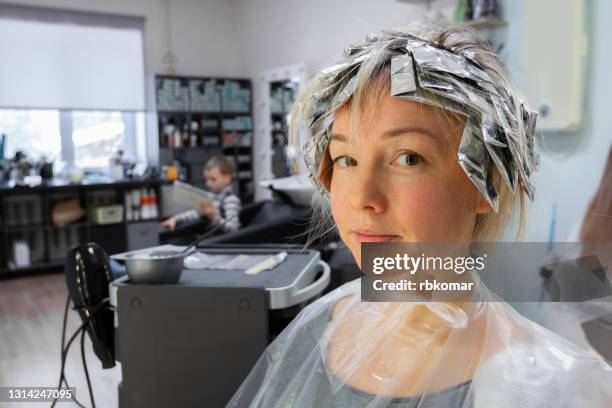 happy young pretty girl sitting in a hairdresser while dyed her hair with foil on her head and looks at the camera. fashion blonde hairstyle creation, bleach, female beauty and hair treatment - blonde hair dye stock pictures, royalty-free photos & images