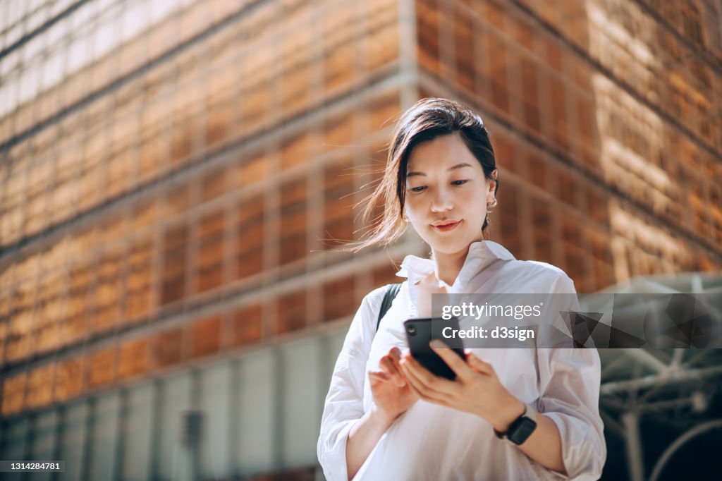 Low angle portrait of young Asian businesswoman checking emails on smartphone outside office building in financial district. With contemporary corporate skyscrapers in background. Making business connections throughout the city